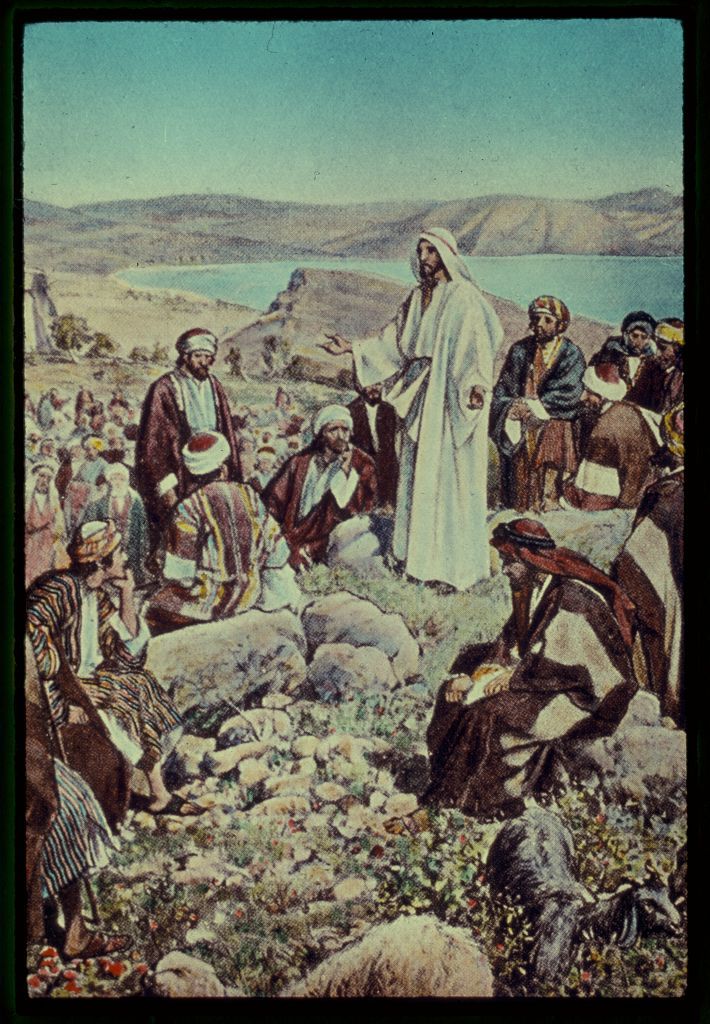 A painting of Jesus standing on a rocky elevated portion of a mountainous region with his arms outstretched speaking to a group of Middle Eastern men during the daytime