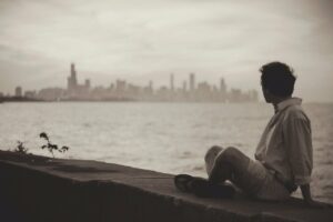 A person sitting on a sea wall in the far right side of the photo looking across a very large body of water towards the skyline of a large distant city