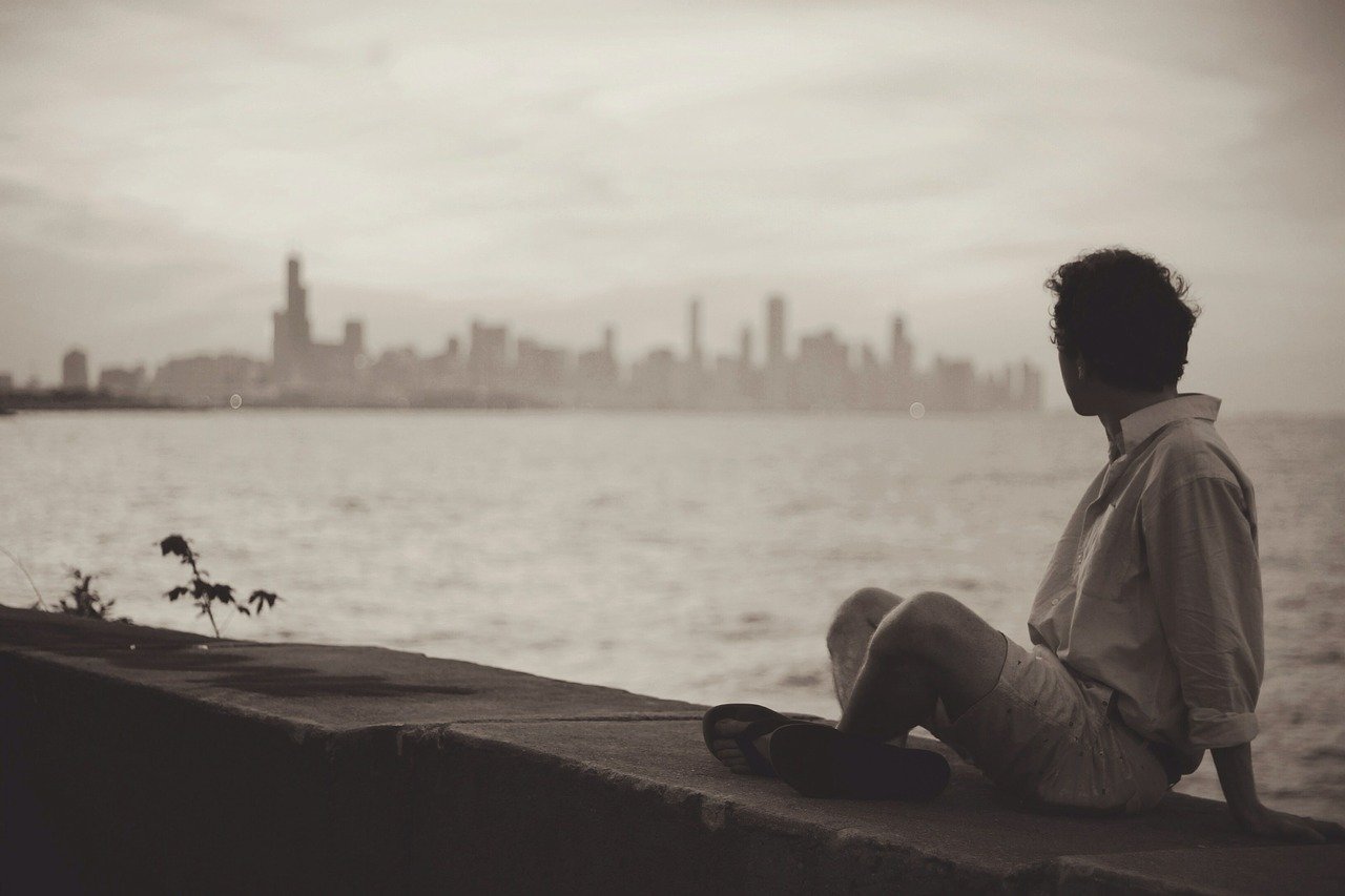 A person sitting on a sea wall in the far right side of the photo looking across a very large body of water towards the skyline of a large distant city
