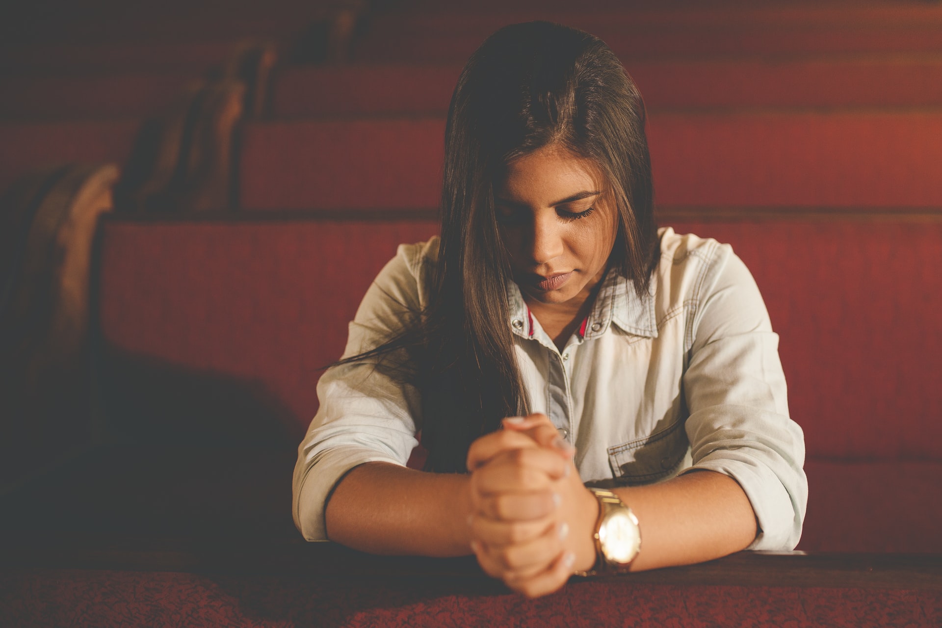A woman with long brown hair sitting in a maroon pew looking down, eyes closed, in prayer with her arms outstretched in front of her and hands clasped together while wearing a large wristwatch with gold band and white face