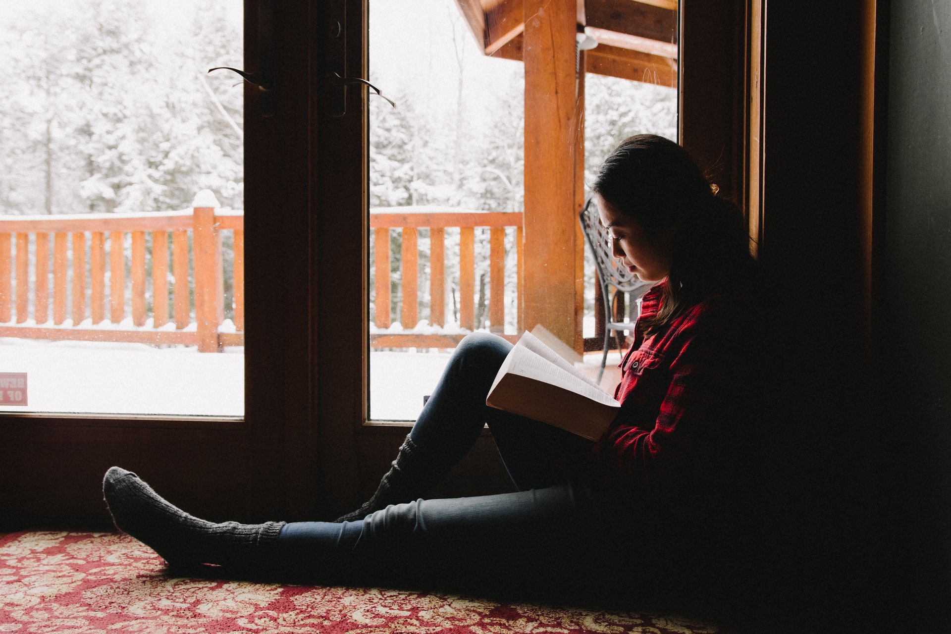 A young woman sitting with one leg outstretched and the other bent to support a book being read while inside a building in front of a pair of doors that open out to a bright, snow covered deck and forest in the background