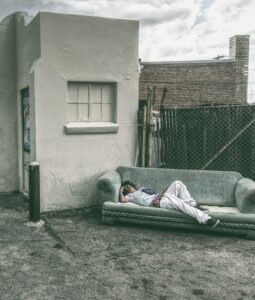 A man in a white shirt and pants sprawled across an old light blue couch asleep with a small white concrete building and painted over window, chain link fence, and a brick building behind