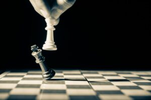 A black and white photo showing a side of view of fingers holding a white chess pawn in the air while knocking down a black chess bishop on a spotlit chess board