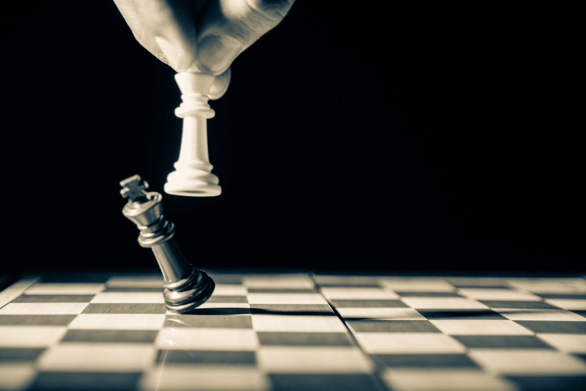 A black and white photo showing a side of view of fingers holding a white chess pawn in the air while knocking down a black chess bishop on a spotlit chess board