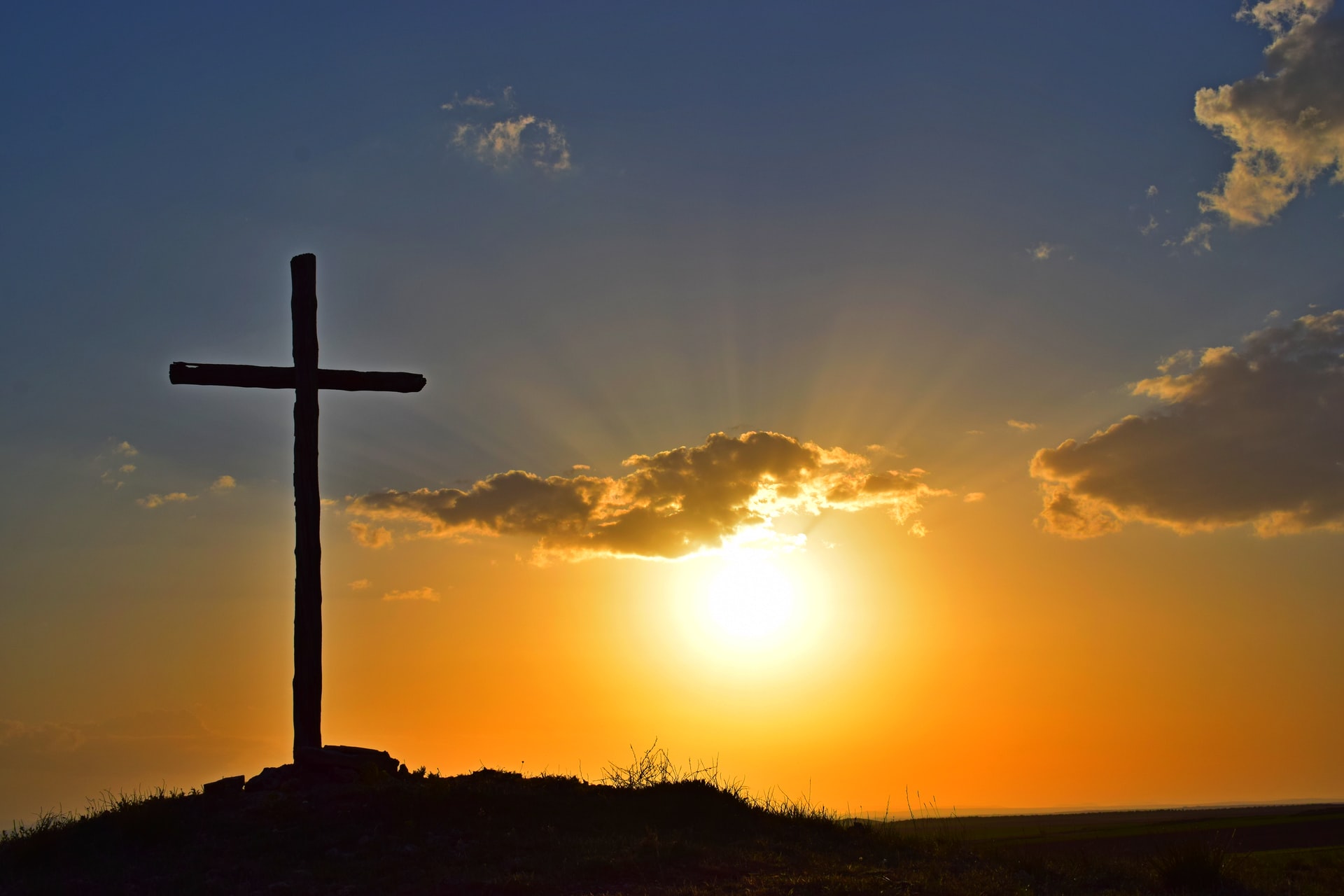A silhouette of a cross on a small hill with sun, clouds and an orange and blue in the background