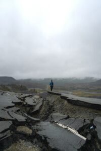 A man with a blue jacket and dark pants walking alongside a collapsed roadway from an earthquake with mountains and heavy fog in the distant background