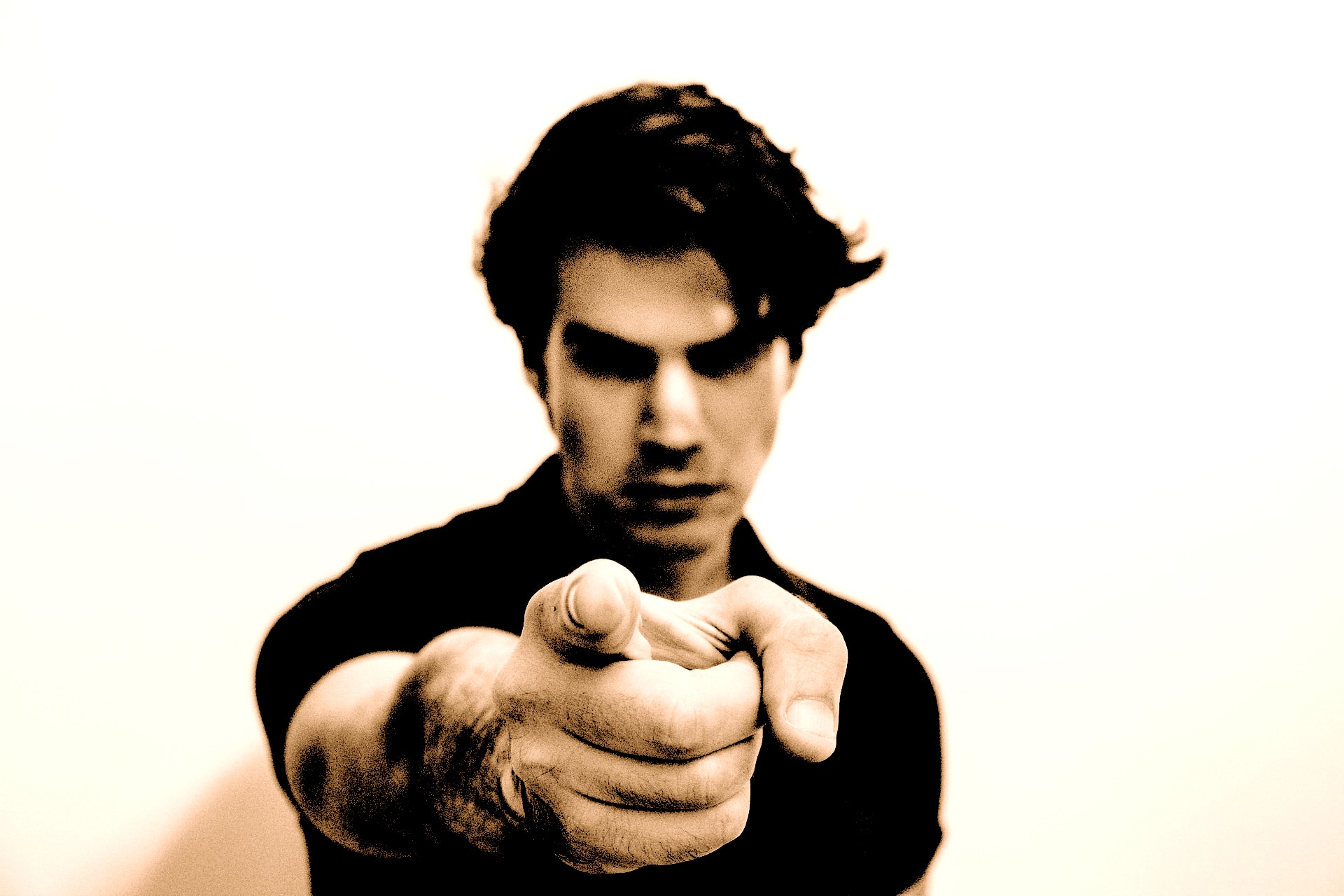 A Caucasian man with dark styled hair and a black shirt looking sternly and pointing index finger at viewer