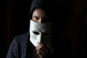 A Caucasian man wearing hooded outerwear while holding a white mask down from his face in a room with light displayed only on his right side