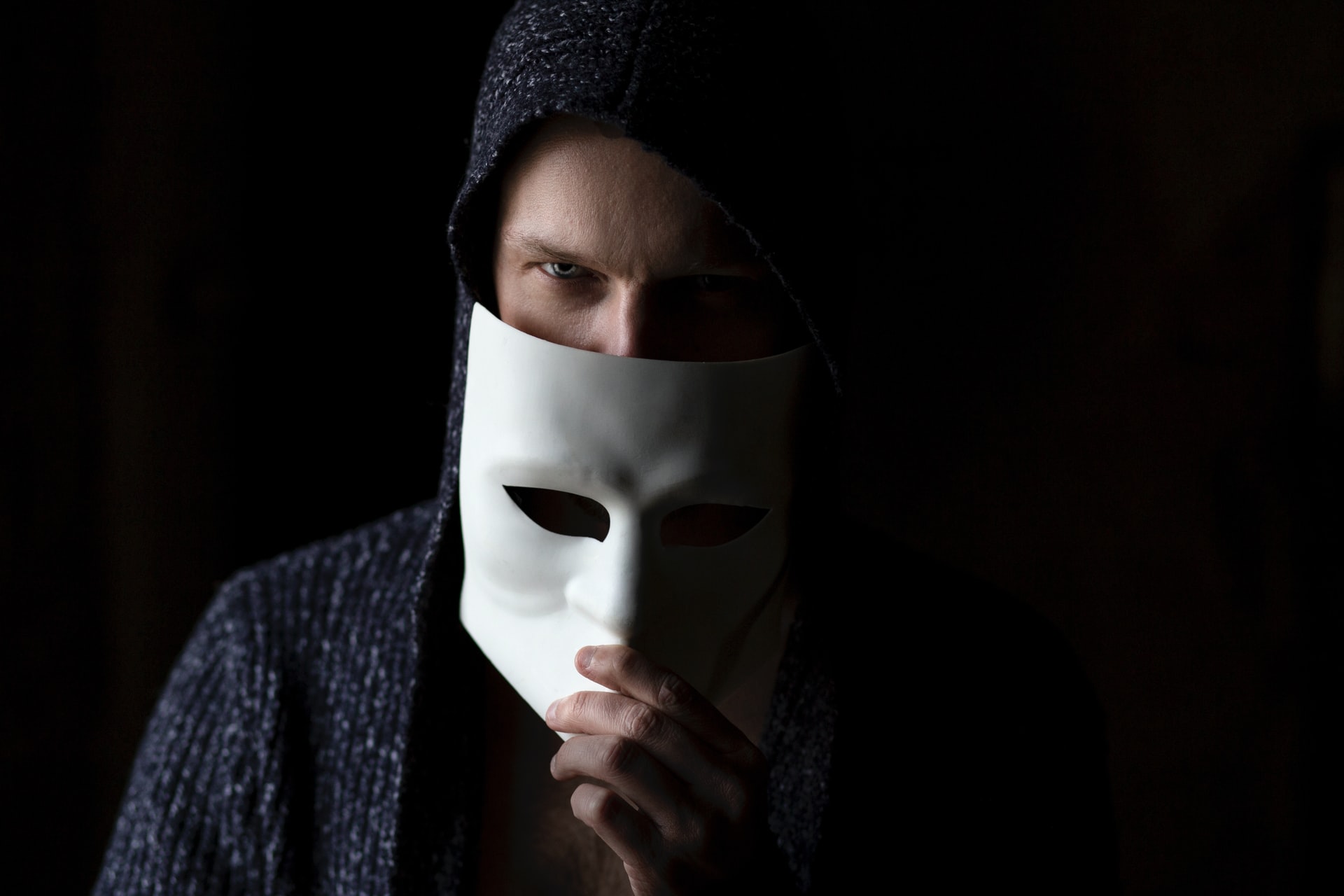 A Caucasian man wearing hooded outerwear while holding a white mask down from his face in a room with light displayed only on his right side