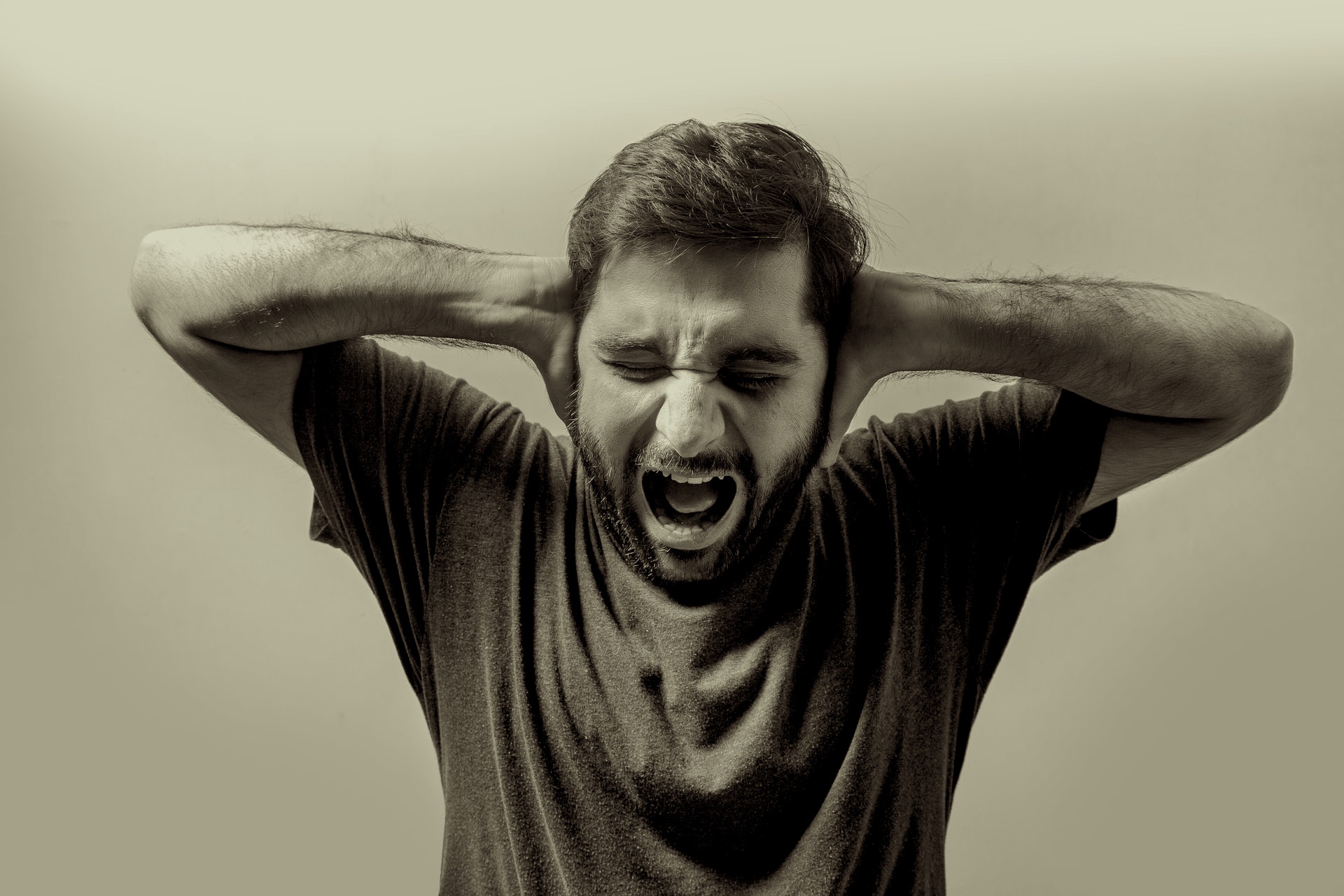 An enhanced black and white photo of a middle aged man with dark hair and beard wearing a dark t-shirt while holding his hands up against his ears and screaming