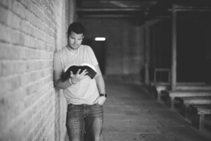Black and white photo of a man standing, leaning against a brick wall while holding an open Bible and looking down in an old rustic church