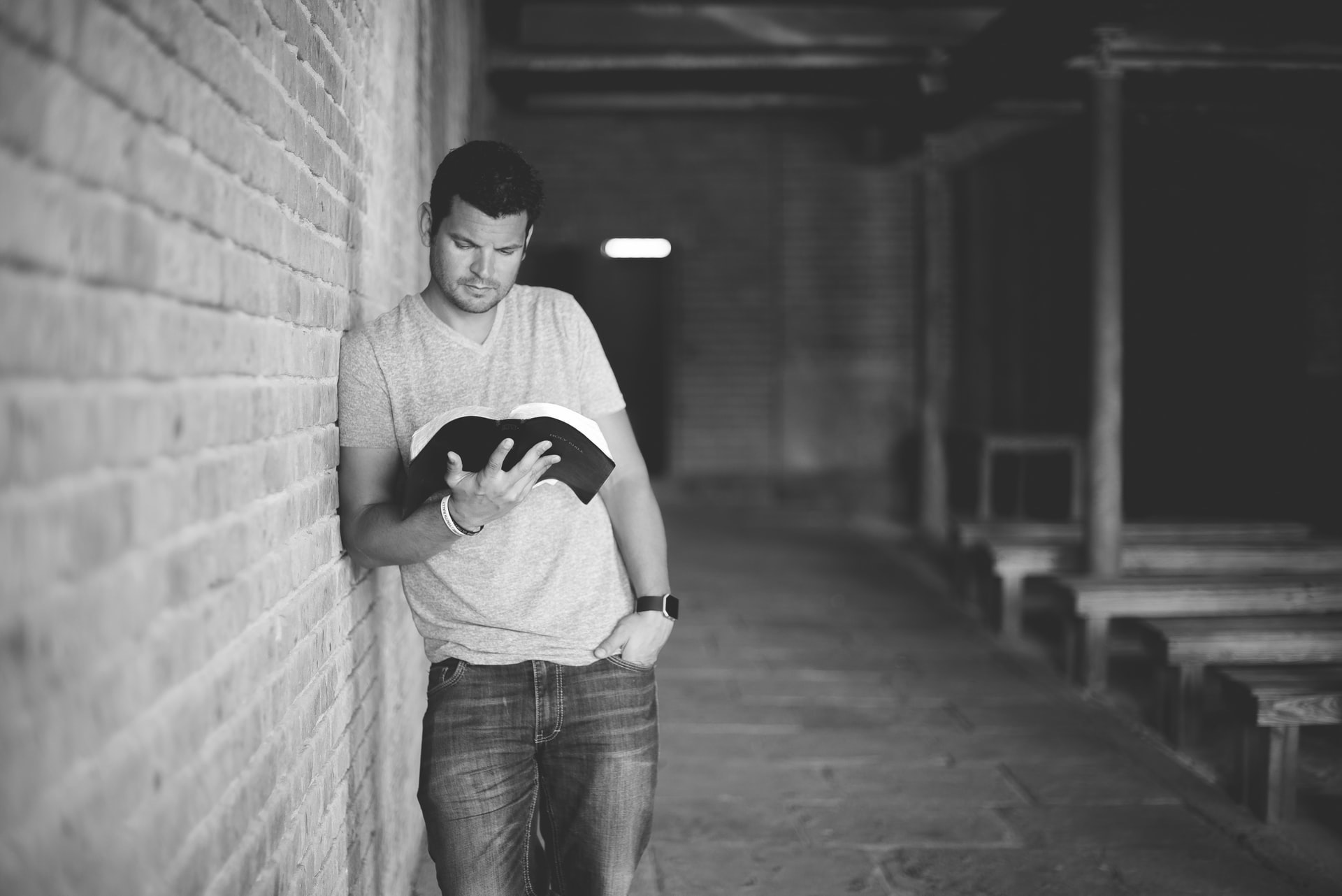 Black and white photo of a man standing, leaning against a brick wall while holding an open Bible and looking down in an old rustic church