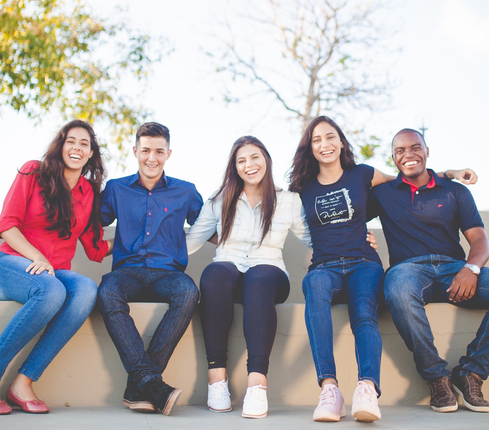 A group of 5 young men and women of mixed races seated in a row and smiling on a sunny afternoon