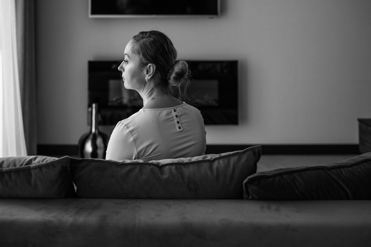 Black and white photo of an older woman wearing a white blouse seated on a sofa looking to a window on the left with an expression of grief