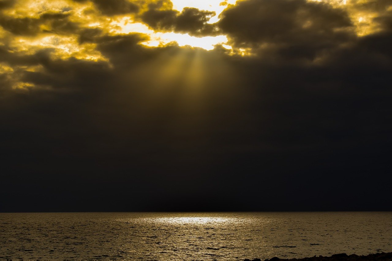 Yellow sunbeams from top of photo streaming down on one area of a large body of water with darkness all around