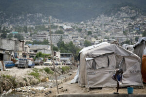 A series of USAID tents with a young man showering in front, several buildings and cars on the left side, and numerous white concrete buildings on a mountainside in the background