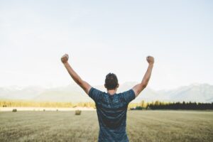 The backside of a Caucasian man with dark hair and a blue and black striped t-shirt holding his clenched fists upward with a harvested hay field and haze covered mountains in the background