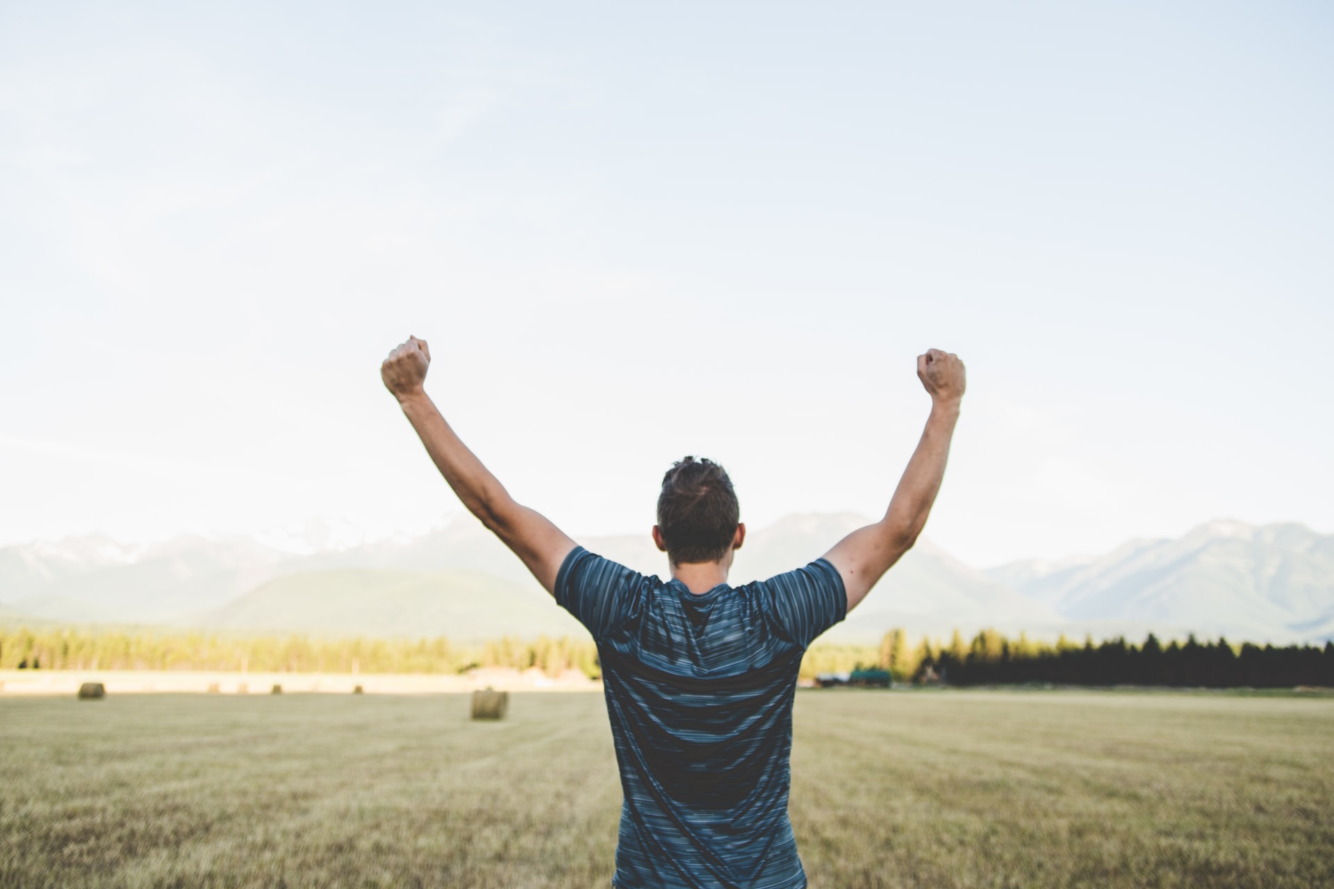 The backside of a Caucasian man with dark hair and a blue and black striped t-shirt holding his clenched fists upward with a harvested hay field and haze covered mountains in the background