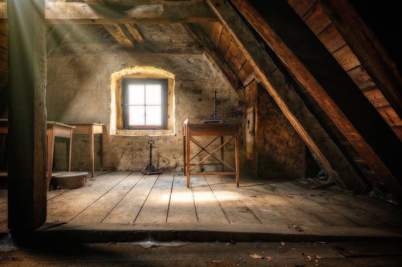 A photo of part of the inside of an old attic with an angled wall, three small tables, a small well-lit window, and sunbeams streaming down from a skylight in the roof