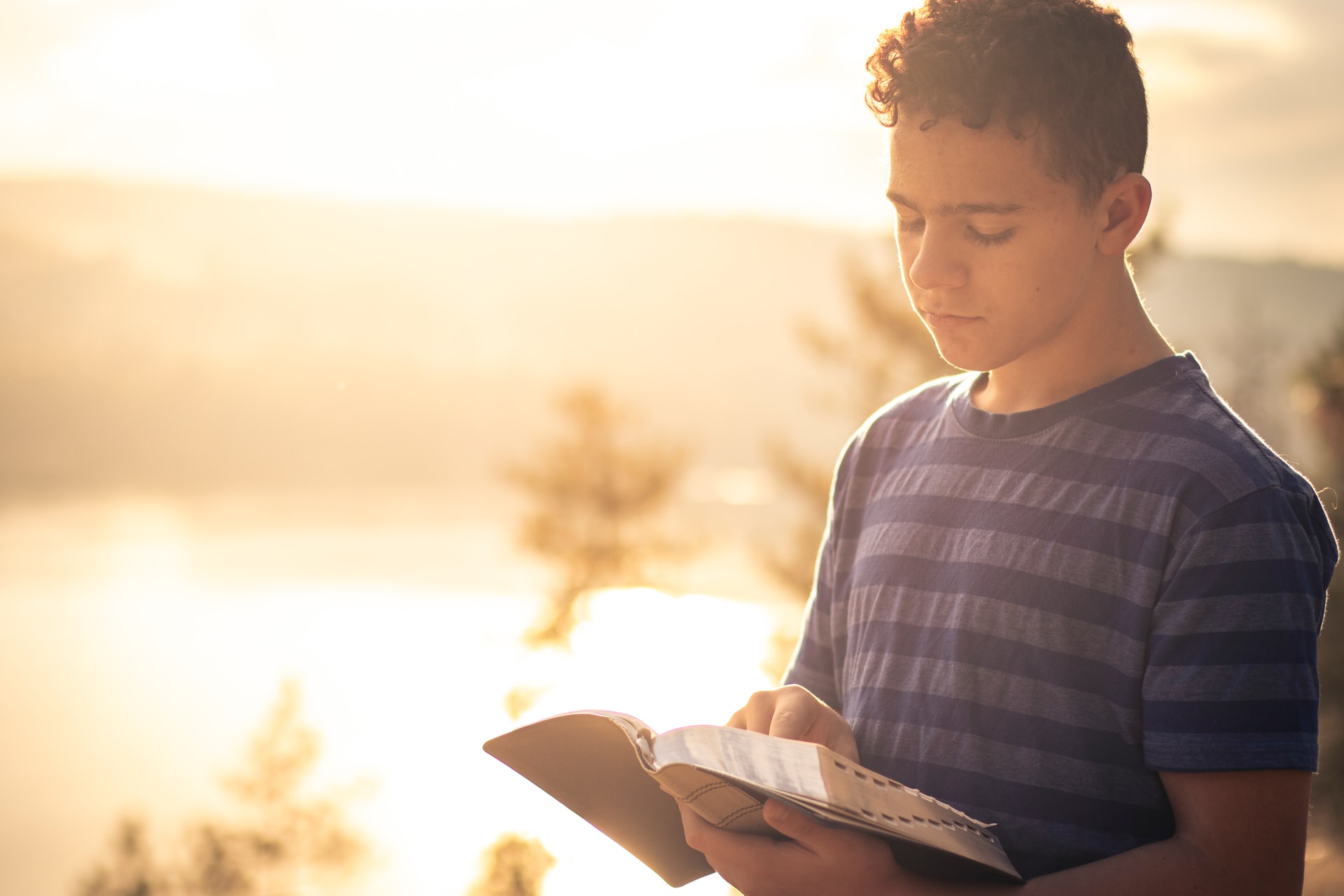 Young male teen wearing a grey and black striped shirt standing and looking down while reading a Bible in his left hand with bright sunlight and a distant mountain in the background