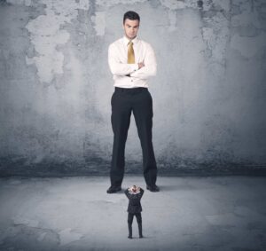 Photo of a very large frowning Caucasian man in a white dress shirt, gold tie and black pants with his arms folded, frowning while looking down at a very tiny man in a black suit with his hands clasped on top of his head in distress