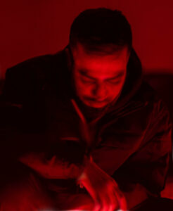 An Asian man with dark hair and wearing a dark jacket seated while leaning forward and looking downward in a red illuminated room