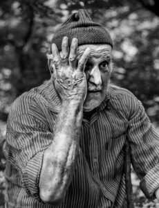 A black and white photo of an older man with a knit hat and a striped short sleeved buttoned shirt seated with his right hand held up against the right side of his face and displaying a stunned expression
