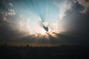 A photo of a partially clouded sky with the beams of an obscured sun shining out in the middle of the picture and a darkened city silhouette below