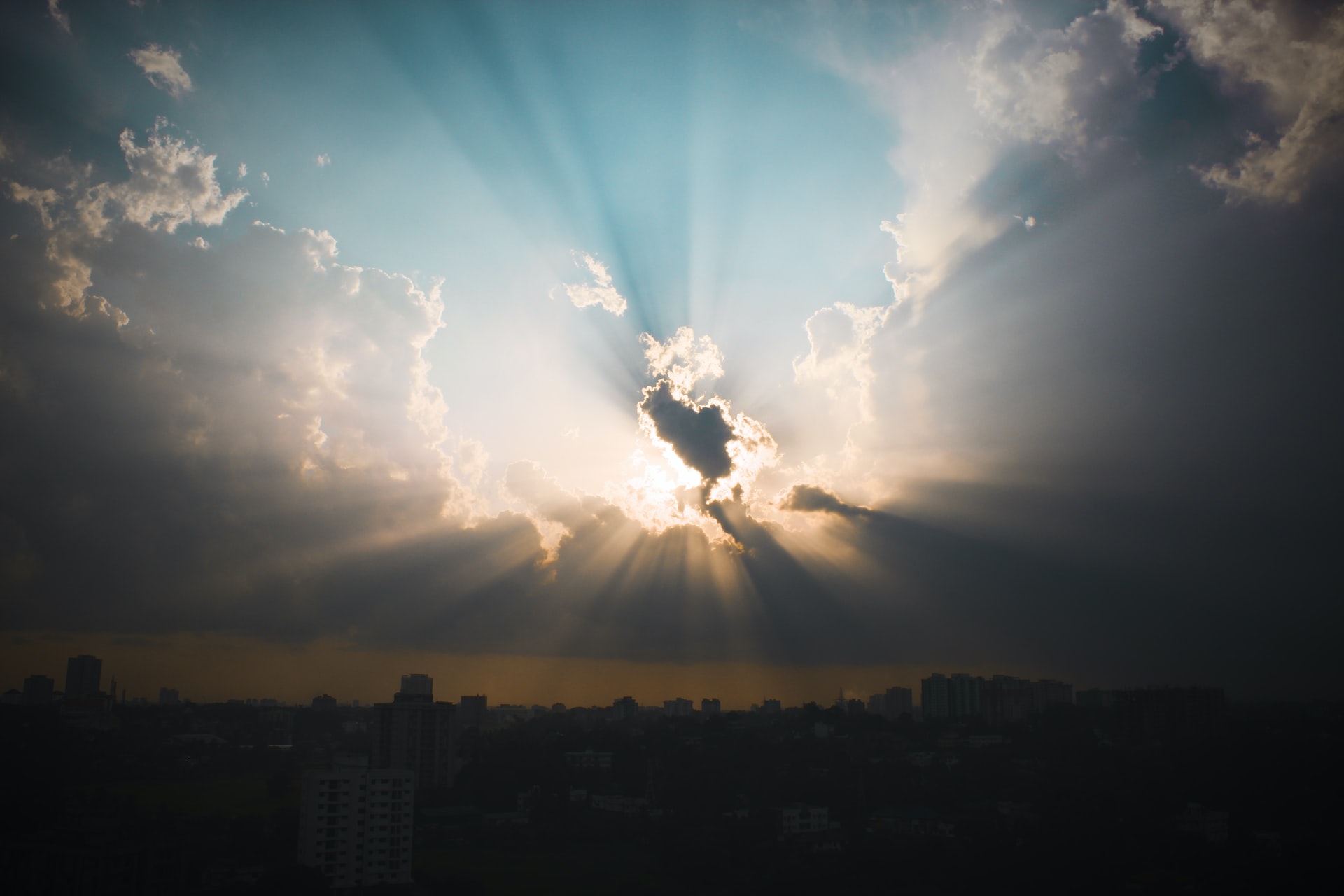 A photo of a partially clouded sky with the beams of an obscured sun shining out in the middle of the picture and a darkened city silhouette below