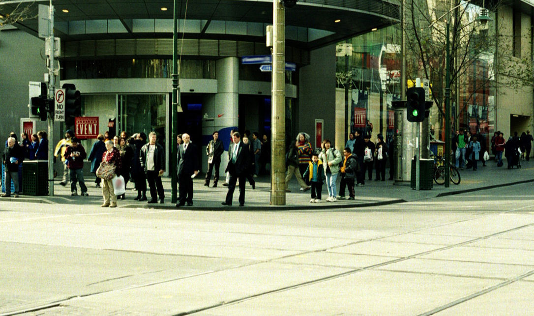 A large group of assorted people on a street corner in front of a department store in a city on a sunny fall day