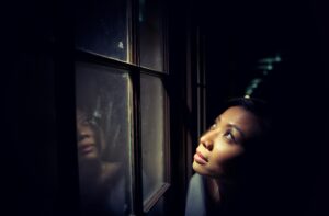 A photo of an Asian woman looking upward outside of a window in a dark room with light shining on her face