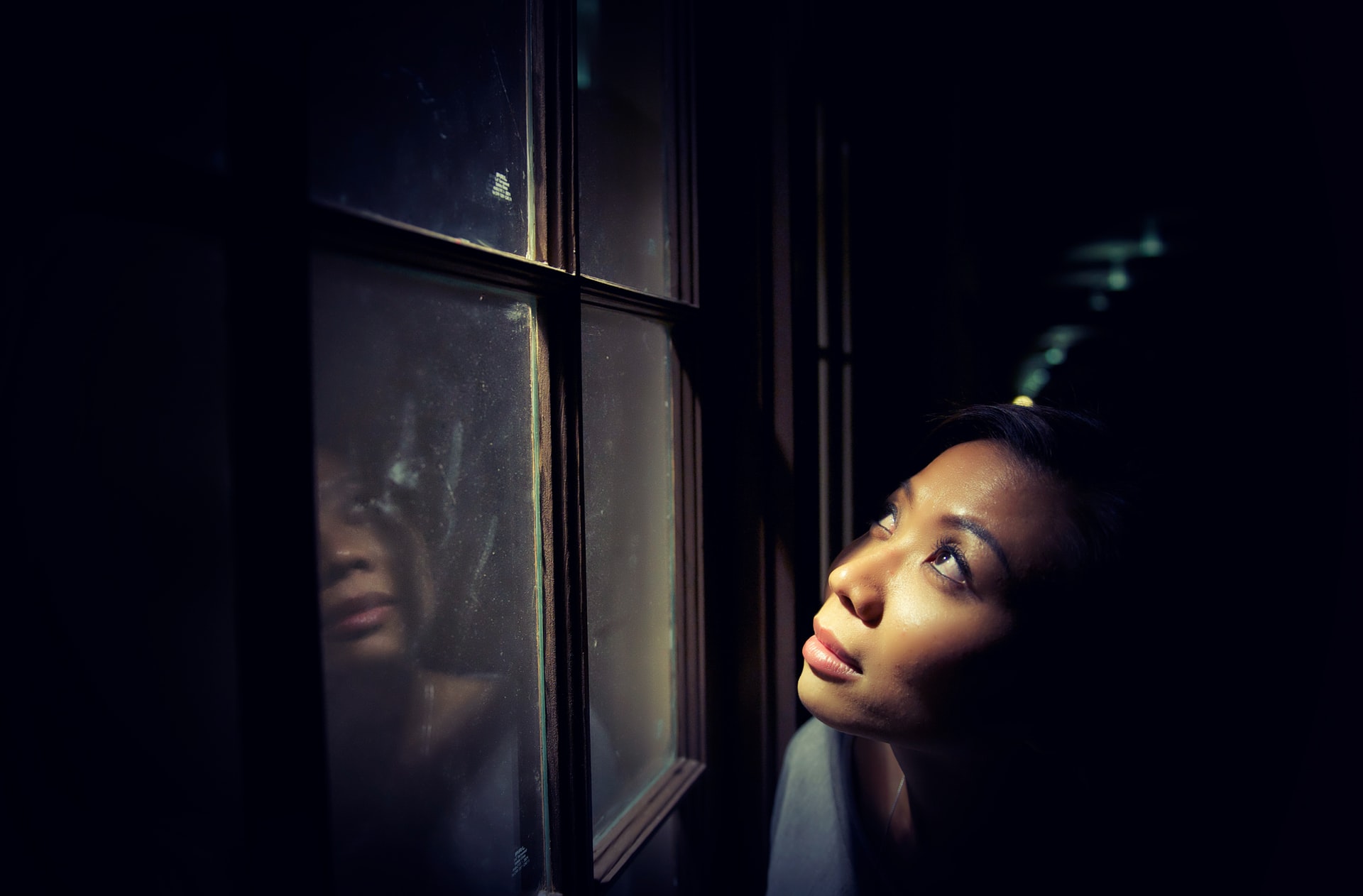 A photo of an Asian woman looking upward outside of a window in a dark room with light shining on her face