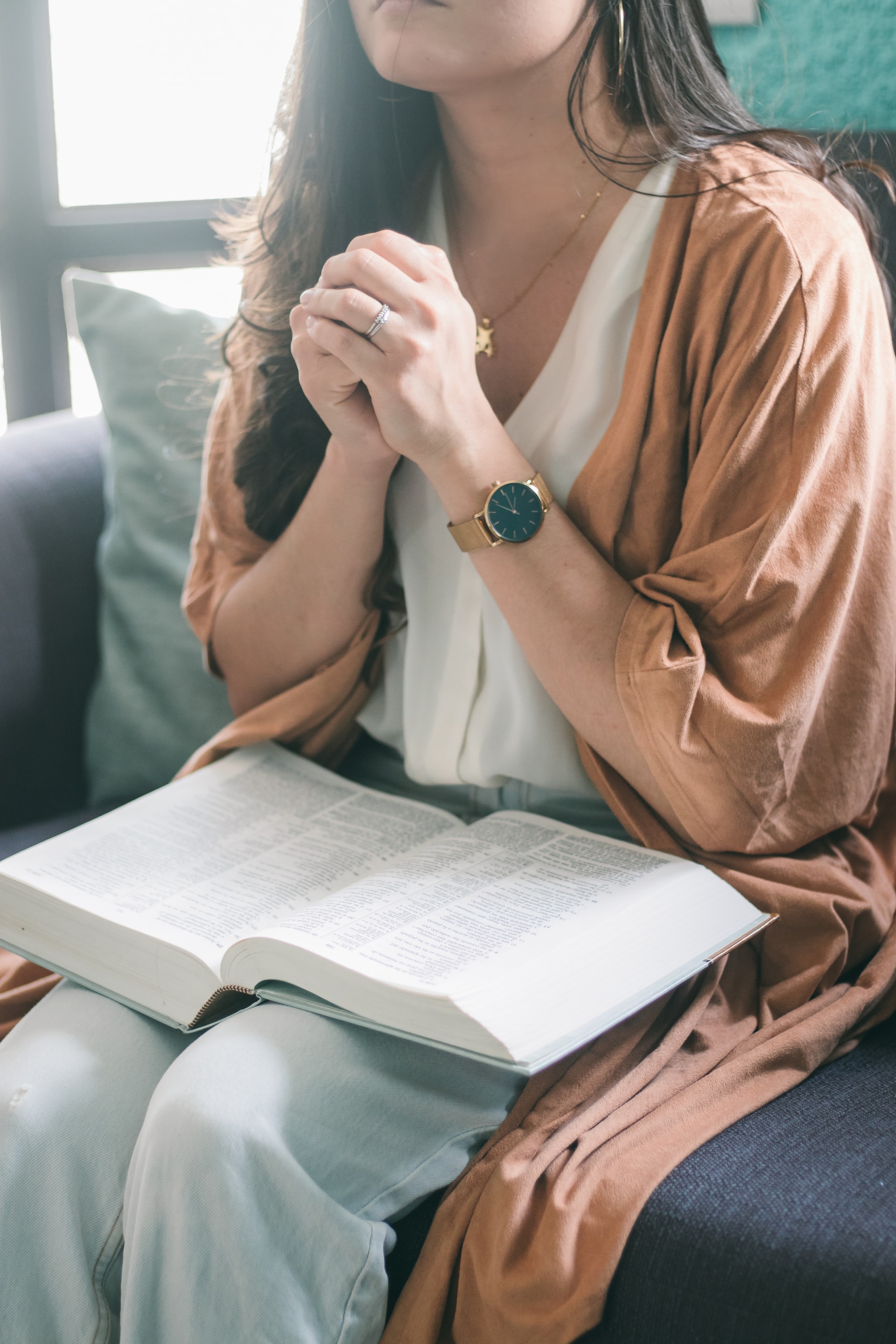 A photo cropped at the neck of a young woman wearing a white shirt, tan cardigan style outerwear, and pale blue jeans with her hands folded together and raised toward her chest while seated in a chair with a Bible on her lap