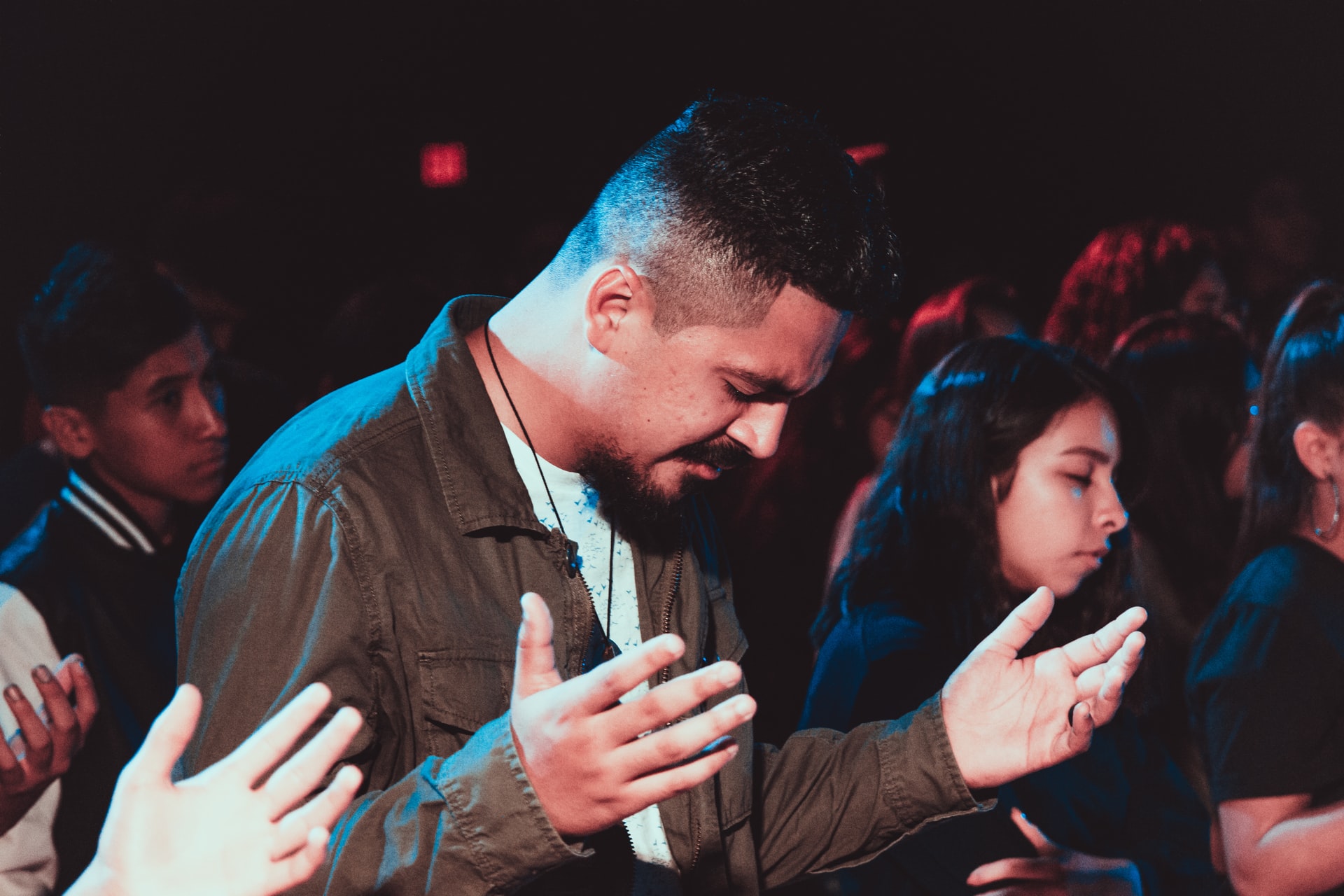 A Hispanic man with his bowed, eyes closed, and hands raised in worship along with others both male and female in the background of a partially darkened room