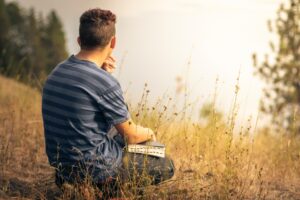 A young Caucasian man wearing a blue and grey striped short sleeved shirt holding a Bible while squatting down in a grassy sloped field with trees and a white sky in the background