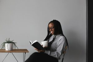 A photo of a young smiling African-American woman wearing a grey sweater and black pants sitting in a chair against a white wall in front of a small wire-framed, wood-topped table supporting a plant while reading a book and holding a white mug