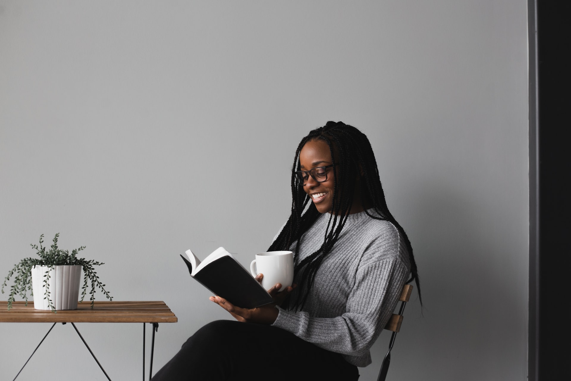A photo of a young smiling African-American woman wearing a grey sweater and black pants sitting in a chair against a white wall in front of a small wire-framed, wood-topped table supporting a plant while reading a book and holding a white mug
