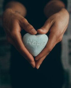 A small, pale blue, heart-shaped stone with the word 'hope' carved in the center held between a pair of outstretched hands also shaped in a 'v' formation