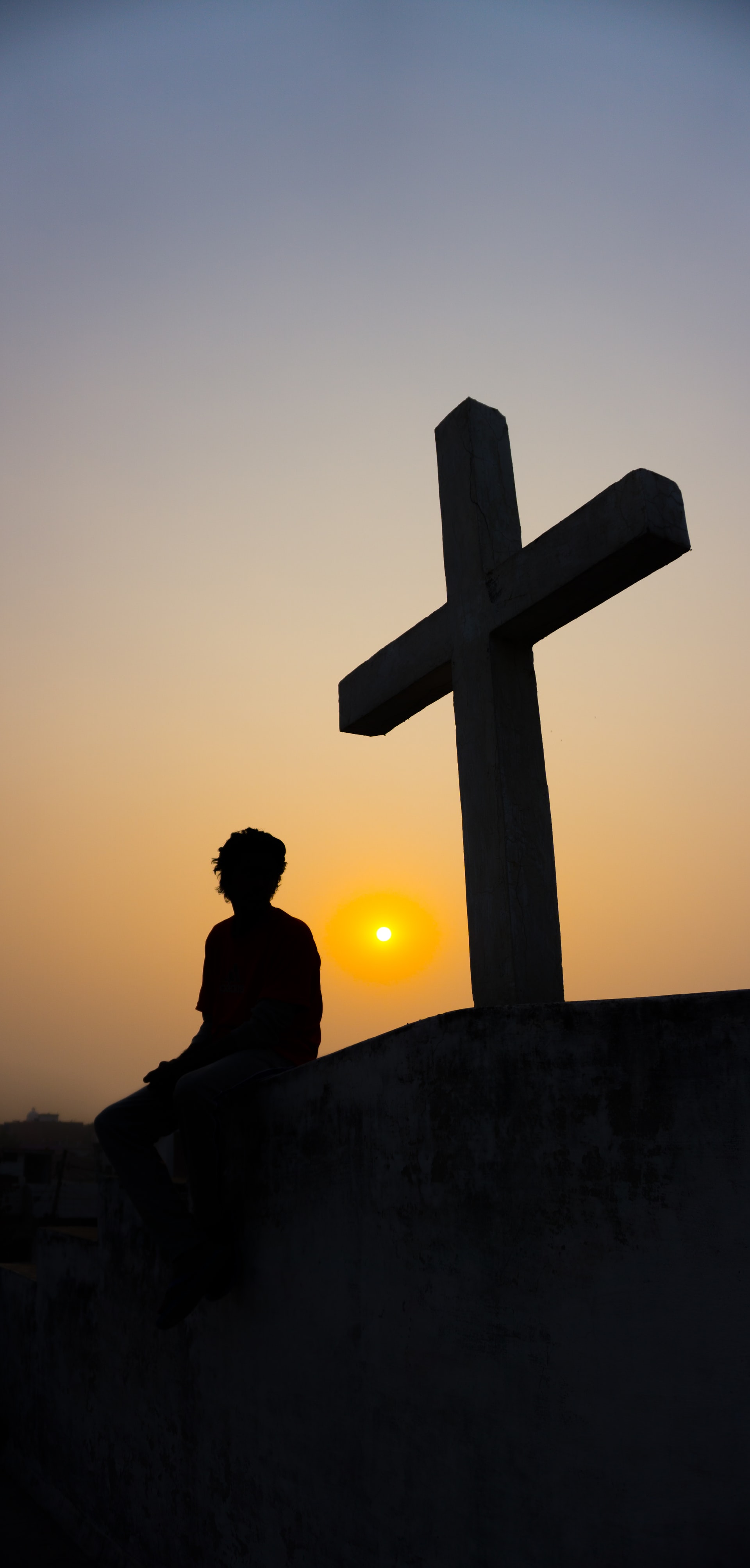 A silhouette of a man seated near a cross on a small hill with a setting sun in the background in a cloudless sky