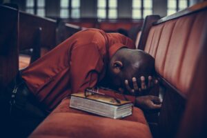 A African man wearing a dark orange shirt and bent down against a pew in a large church with his hands against his face in prayer with a Bible and a pair of glasses beside him