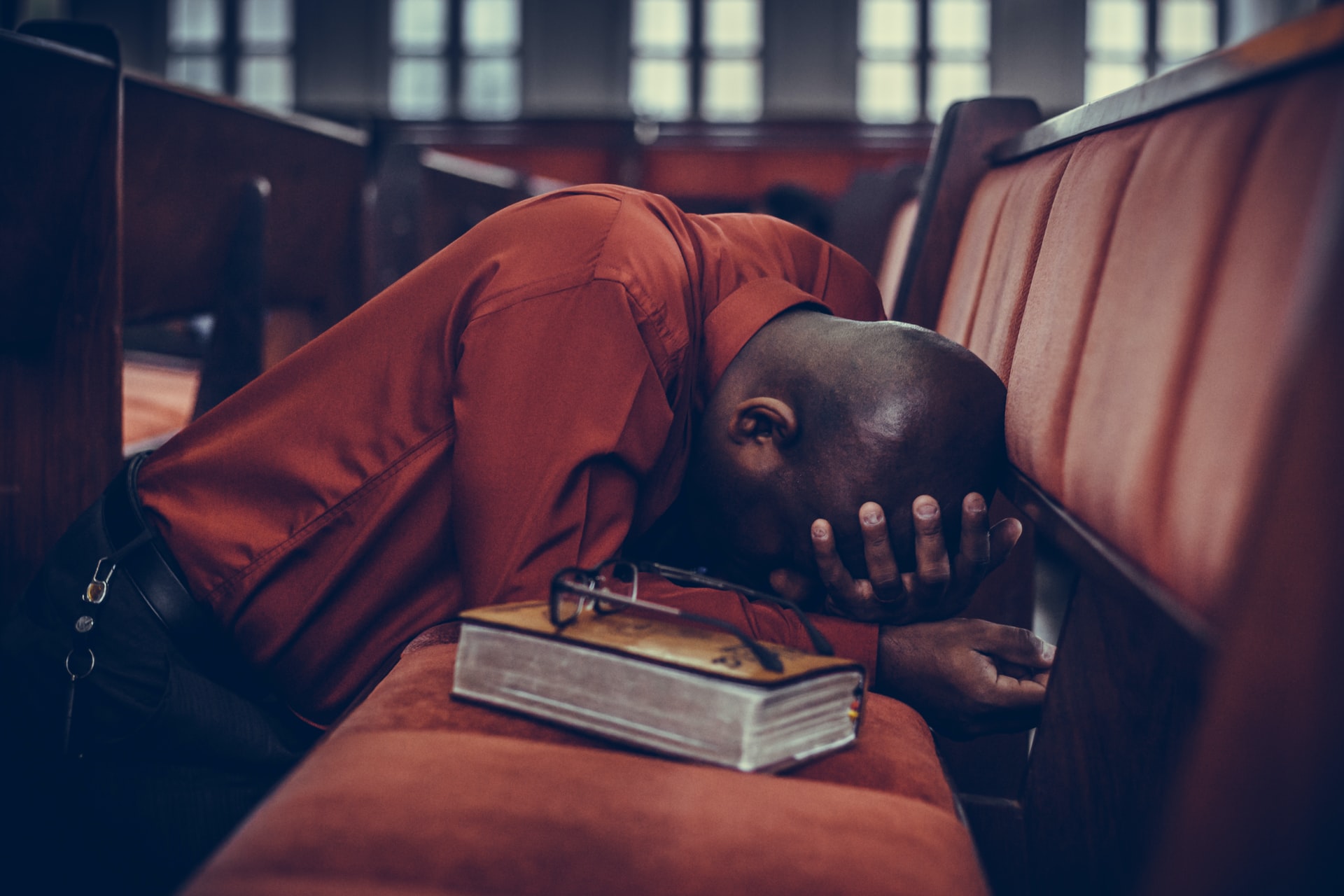 A African man wearing a dark orange shirt and bent down against a pew in a large church with his hands against his face in prayer with a Bible and a pair of glasses beside him