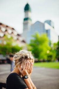 A blond haired Caucasian woman seated with her hands pressed against and obscuring her face in the midst of a urban park with a blurred background