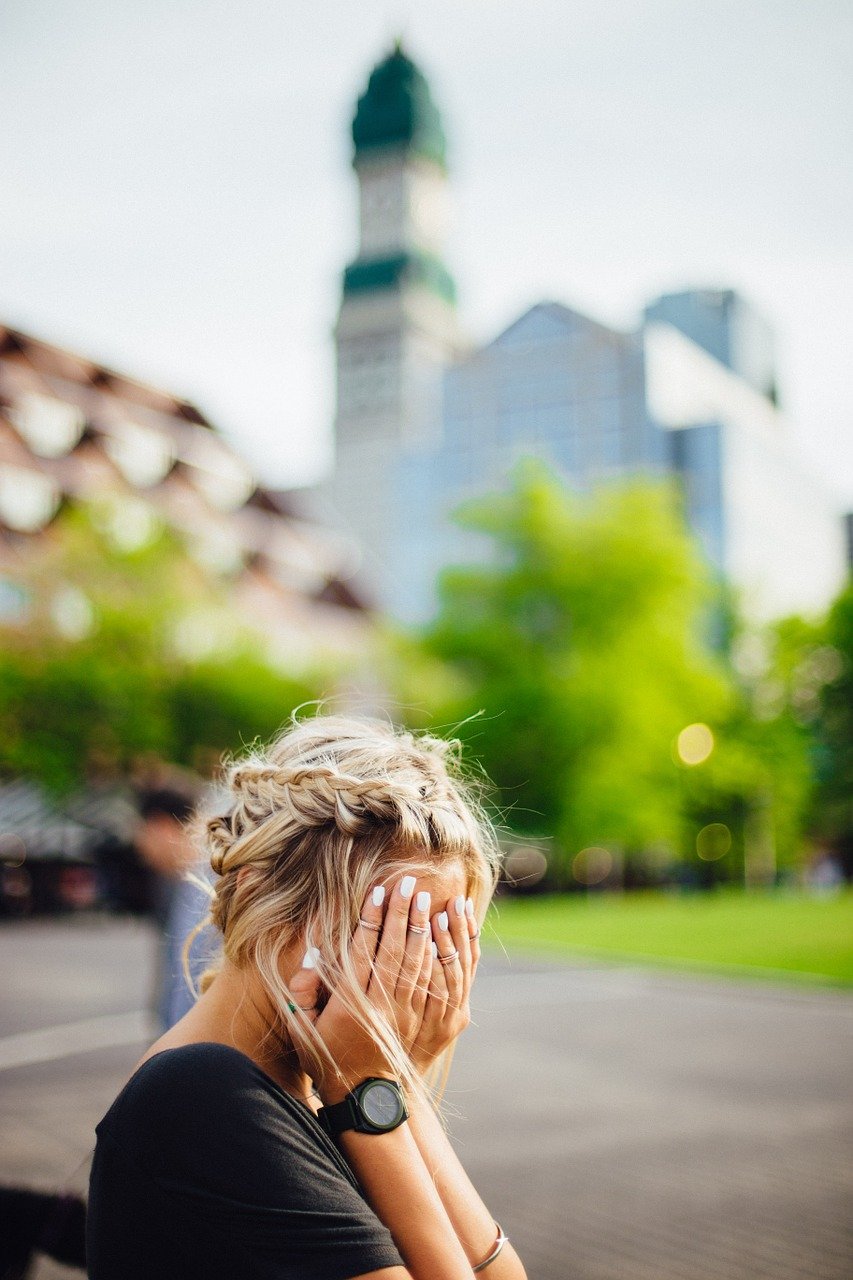 A blond haired Caucasian woman seated with her hands pressed against and obscuring her face in the midst of a urban park with a blurred background
