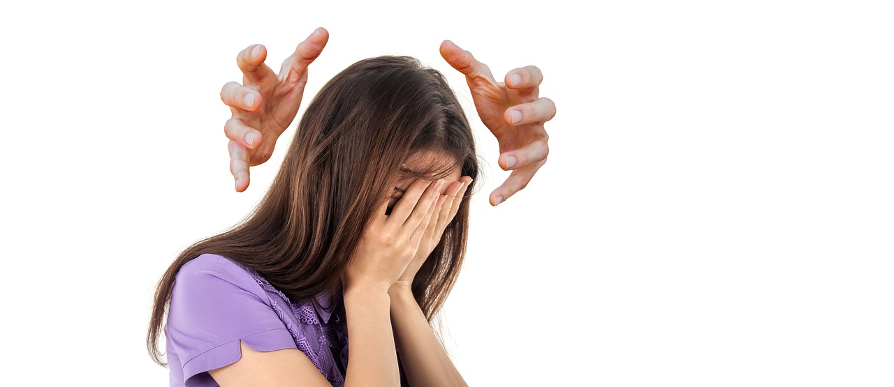 A young Caucasian woman with long straight brown hair wearing a short sleeved lavender colored shirt intimidated by a power greater than herself while holding her hands up against her face while a pair of disembodied hands surround her head in an attempted attack
