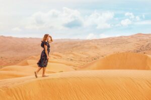 A young Caucasian woman wearing a dark flower print dress while walking on a dune in a desert with her left hand held up to her eyes as she looks forward and hazy clouds on a blue sky in the background