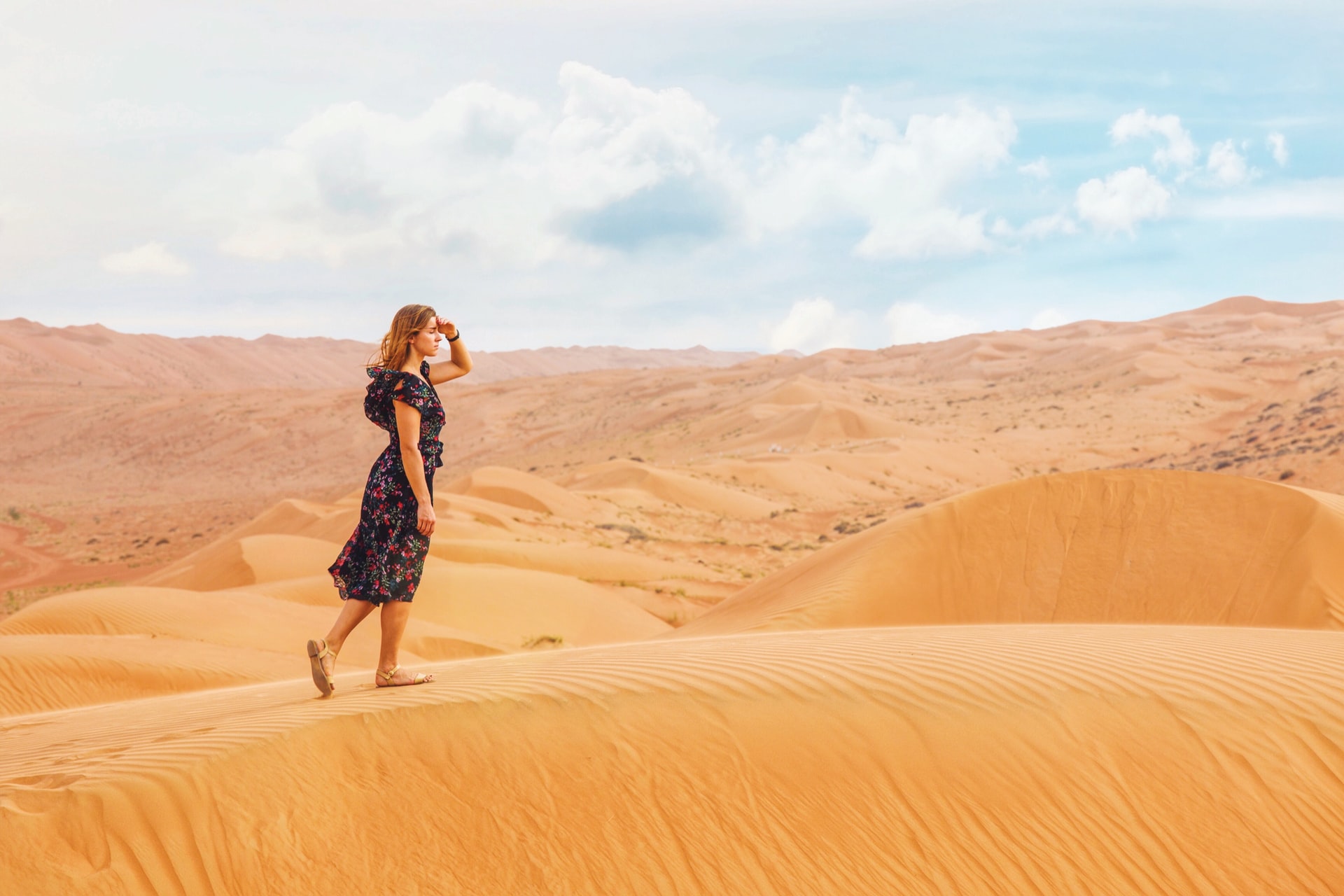 A young Caucasian woman wearing a dark flower print dress while walking on a dune in a desert with her left hand held up to her eyes as she looks forward in hope with hazy clouds on a blue sky in the background