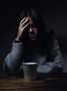 A young African or Asian woman wearing a grey sweater seated at a table in a darkened room with a coffee cup in front and her right hand held up against her forehead and her eyes closed