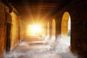 An exterior hallway lined with stone walls, archways, and a partially covered fog floor ending with a brightly streaming sun