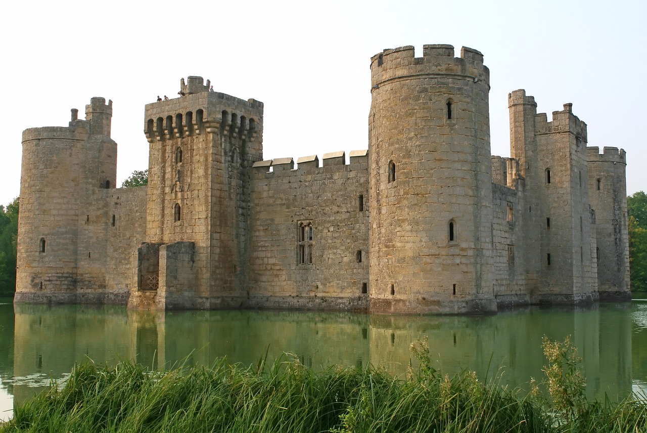 A large stone castle sitting in the middle of a green pond with grasses in the foreground and a pale sky overhead