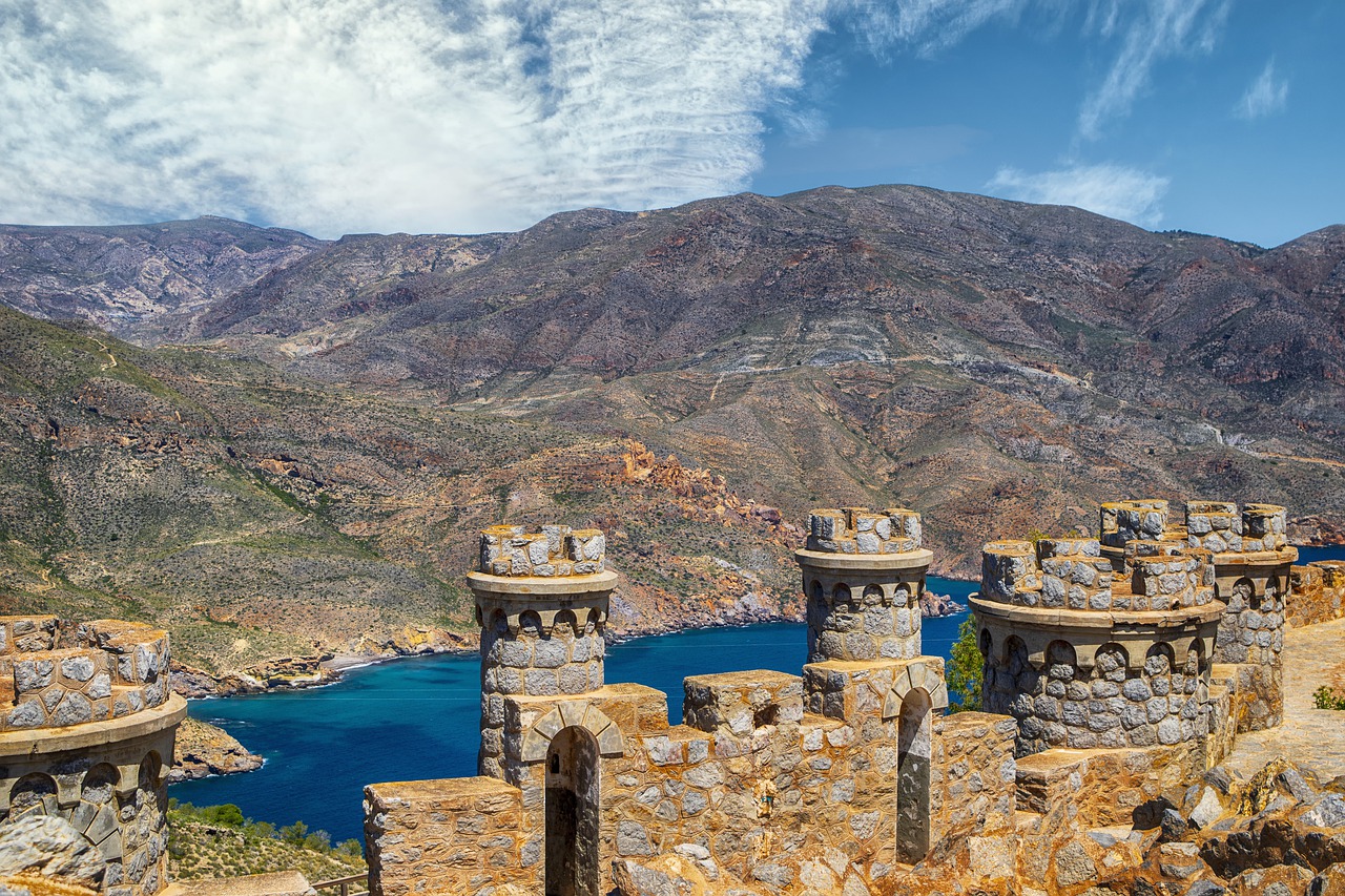 Looking out across the top of a stone castle surrounded by mountains, dark blue water, and a partially clouded blue sky